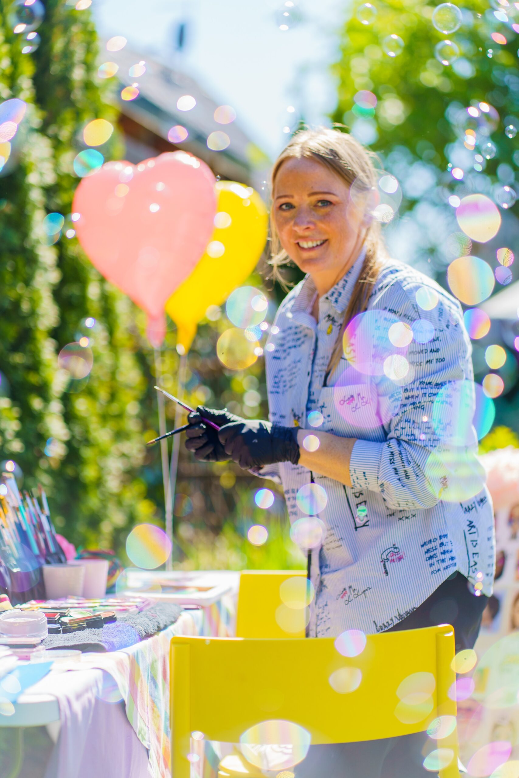 Daniela Staigys steht lächelnd inmitten von bunten Luftballons und Wassermalfarben vor einem gelben Stuhl.