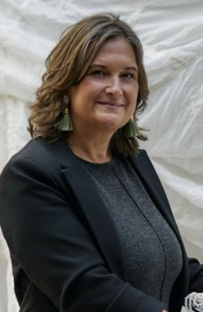 Antonia Neubaur lächelt in die Kamera - sie hat mittellange gelockte brauche Haare und trägt auffällige grüne Ohrringe zu einem schwarzen Blazer und grauem Oberteil.