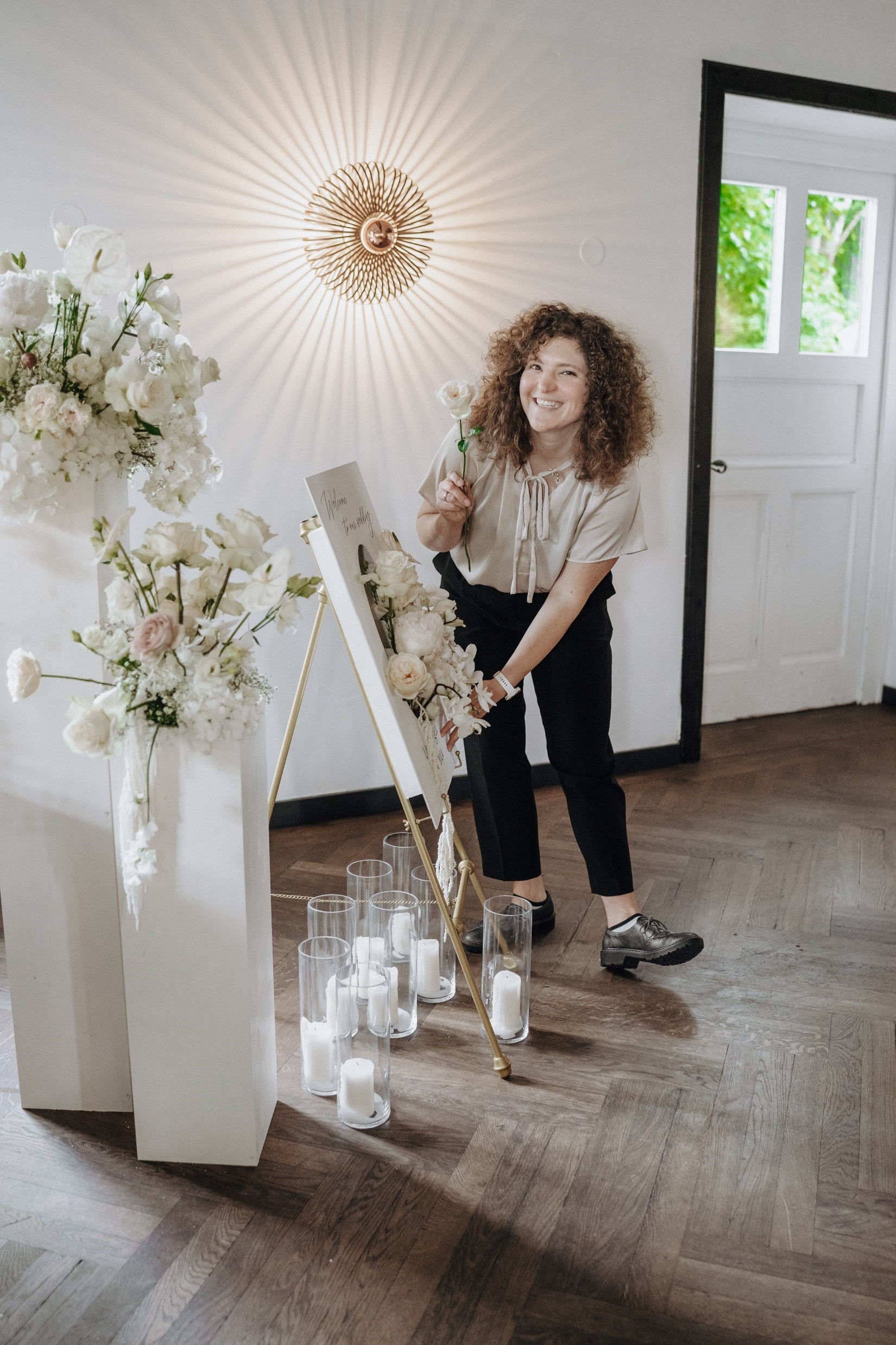 Julia Helbig lacht herzlich und steht vor einem Blumen- und Kerzenarrangement mit Leinwand in weiß.