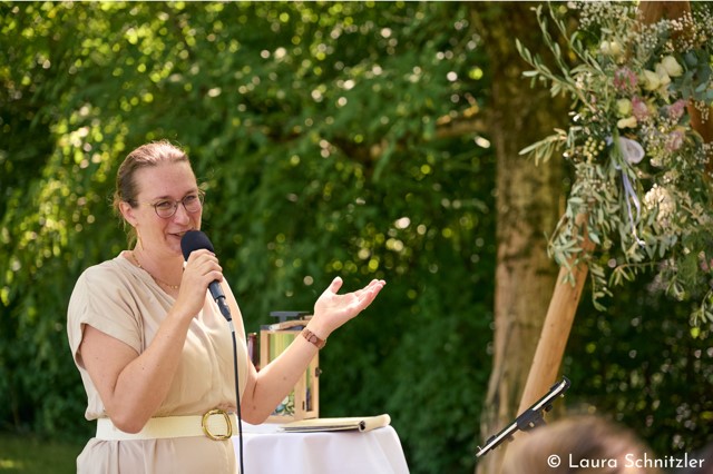 Die freie Rednerin Theresa Henckell spricht freudestrahlend über Mirkophon. Es ist ein gSommertag, im Hintergrund üppige grüne Büsche und Bäume. 